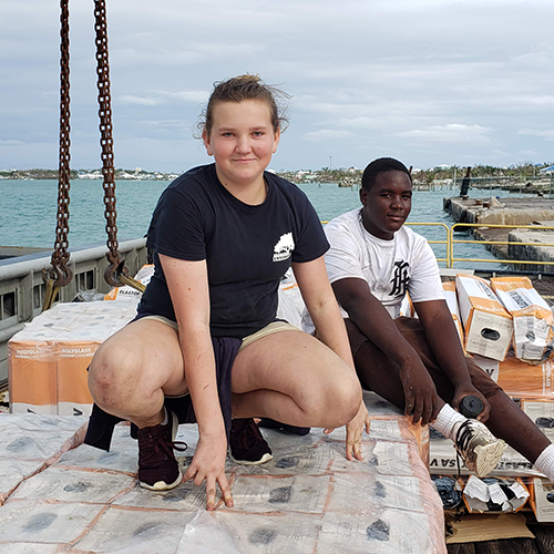 Teenagers pose on top of pallets of roof underlayment donated by Polyglass to the disaster relief efforts in the Bahamas.