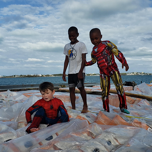 Three boys in superhero costumes play on top of pallets of roof underlayment on the back of a shipping boat in the Bahamas.