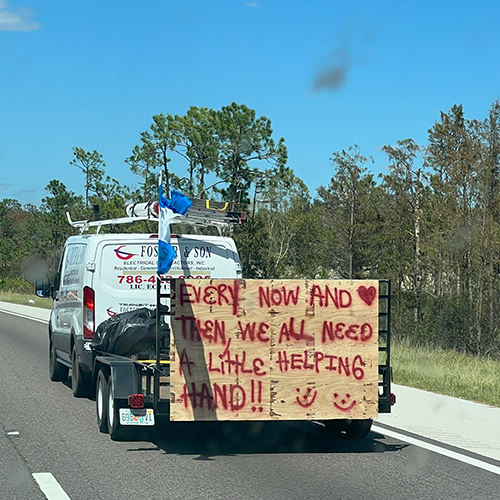 Van pulling a trailer with a painted sign that reads "Every now and then we all need a helping hand!"