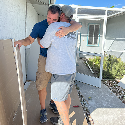 Two men hugging while prepping drywall to be hung inside a widow's home.