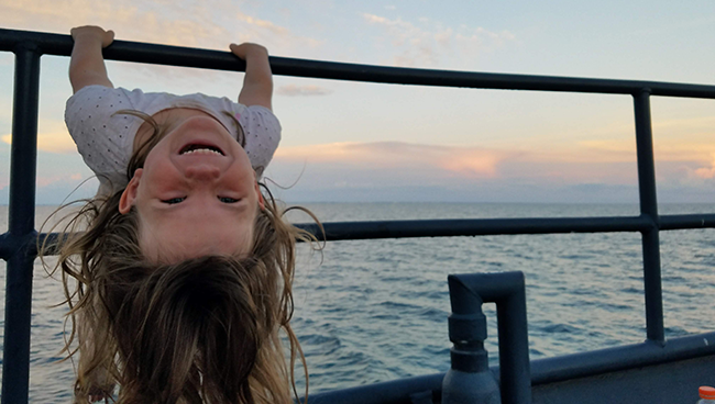 Little girl hanging upside down from the rails of a boat out on the ocean at sunset.