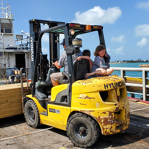 Little girl rides on the back of a forklift while her dad unloads donated lumber during hurricane recovery efforts.