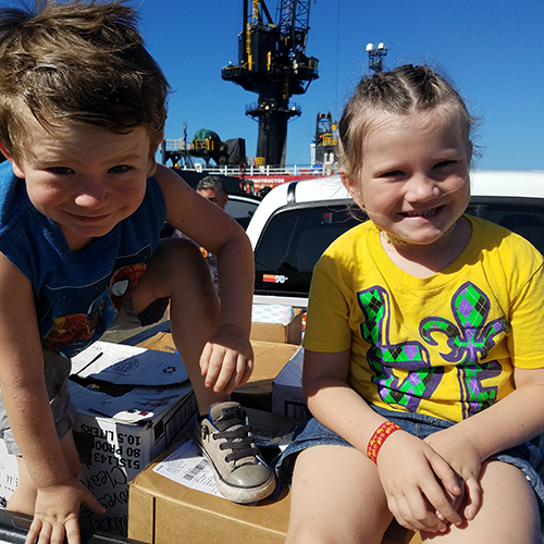 Two smiling children sit on a pile of donations in the bed of a truck on their way to distribute food and water to those effected by Hurricane Maria.